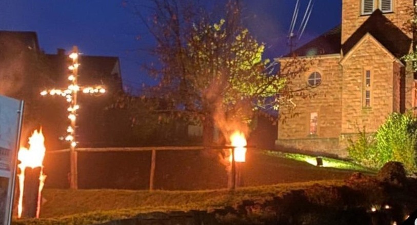 Ein brennendes Kreuz und Fackeln werden nachts vor einem Gebäude angezündet, wobei die Flammen die Umgebung beleuchten.