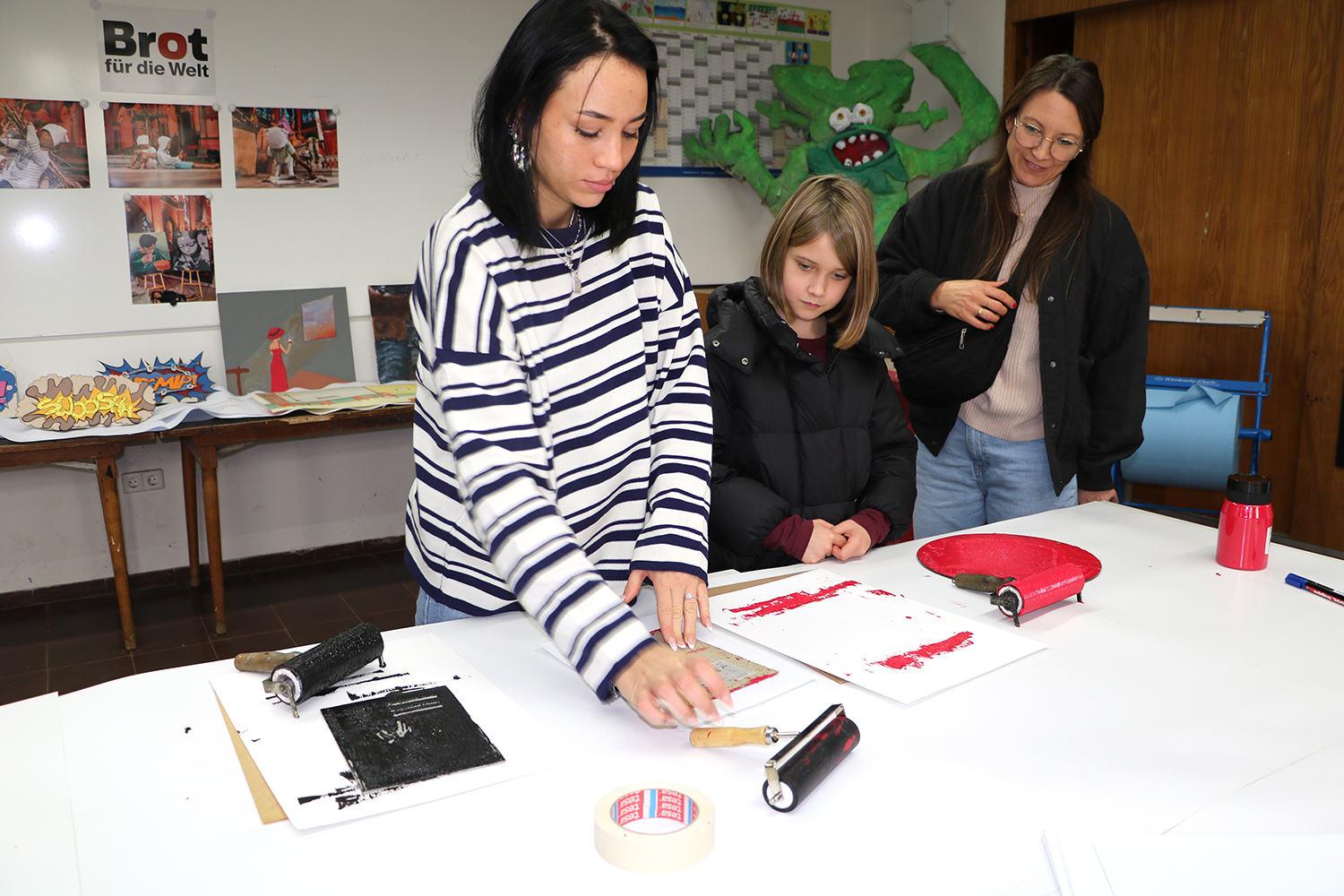 Drei Personen verwenden Farbrollen und Papier für ein Kunstprojekt an einem Tisch in einem kreativen Klassenzimmer.