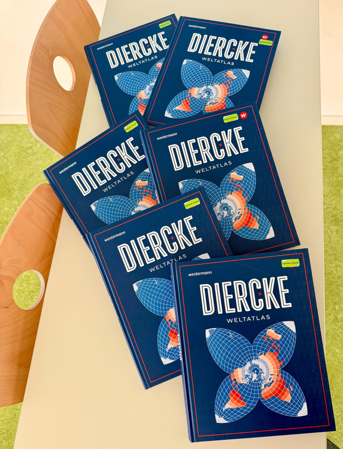 Sechs blaue Diercke-Weltatlanten liegen auf einem hellen Tisch neben einem Holzstuhl.