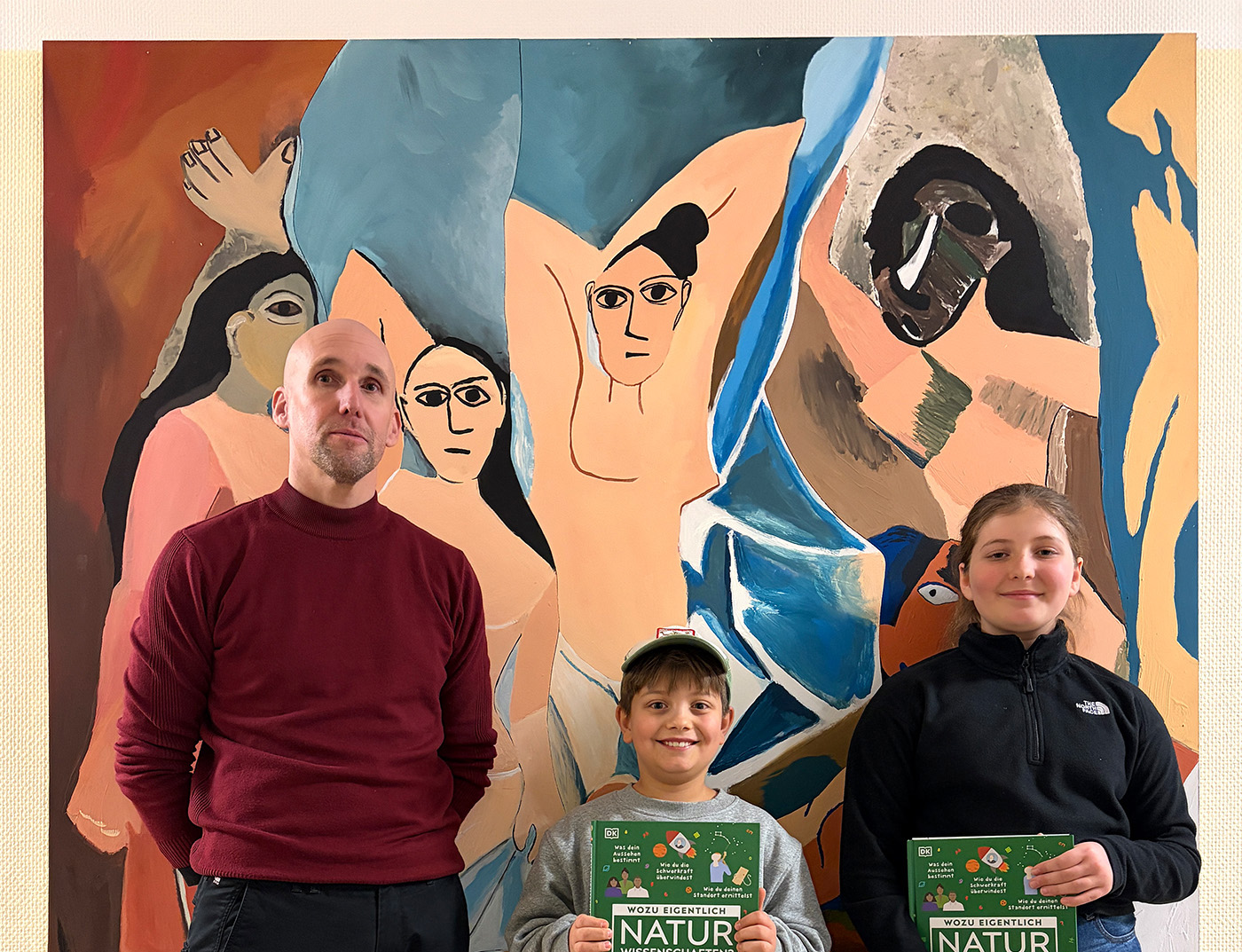 Ein Erwachsener und zwei Kinder stehen lächelnd vor einem farbenfrohen abstrakten Wandgemälde und halten Bücher mit dem Titel "NATUR.