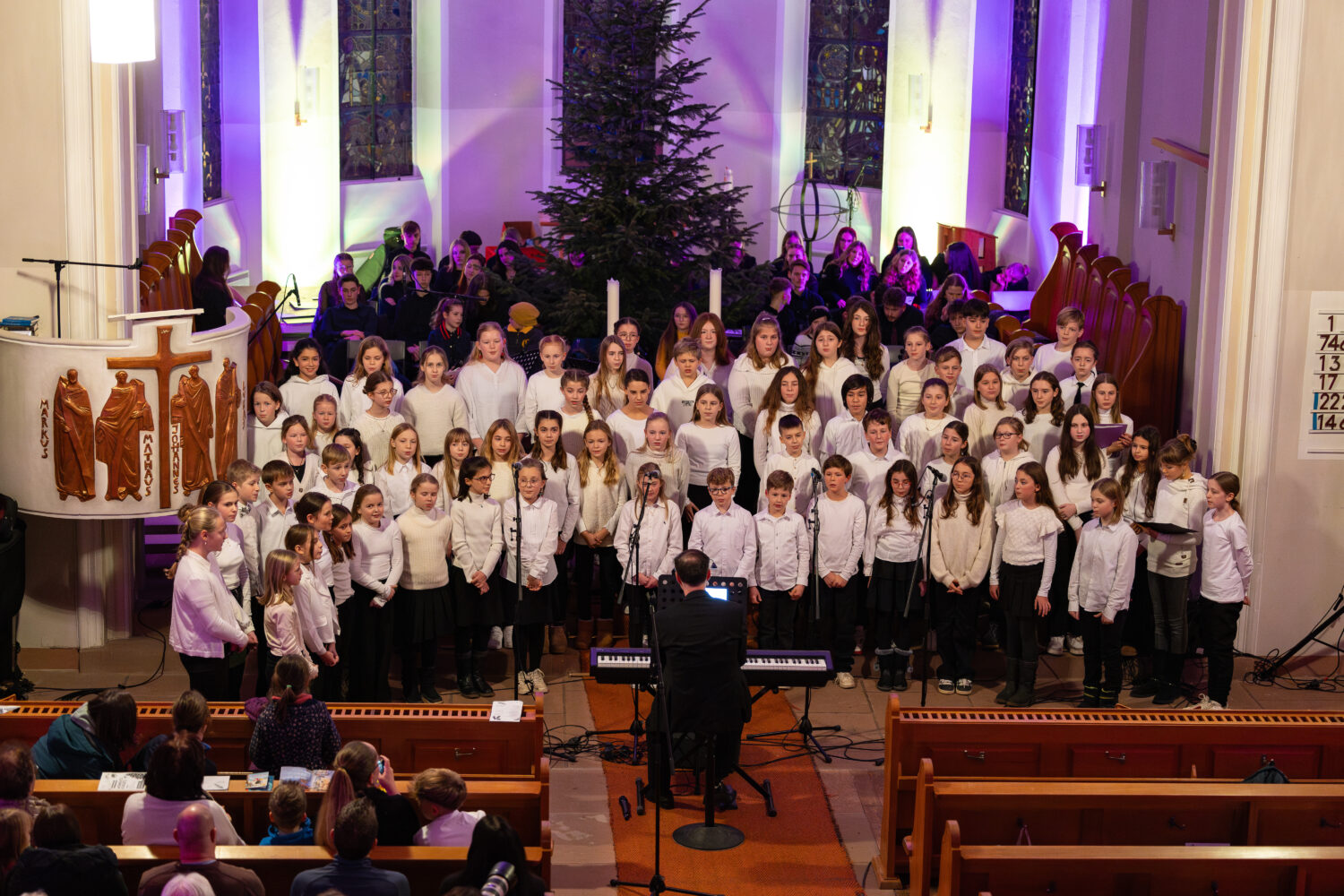 Ein Kinderchor singt in einer Kirche unter der Leitung eines Dirigenten vor einem Publikum, das in den Kirchenbänken sitzt.