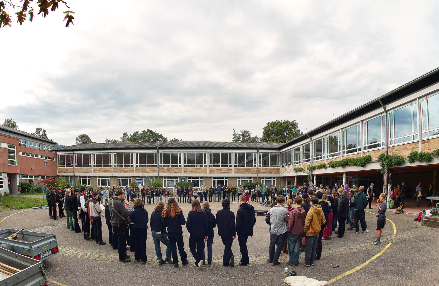 Eine große Gruppe von Menschen bildet einen Kreis auf einem Schulhof unter einem bewölkten Himmel.