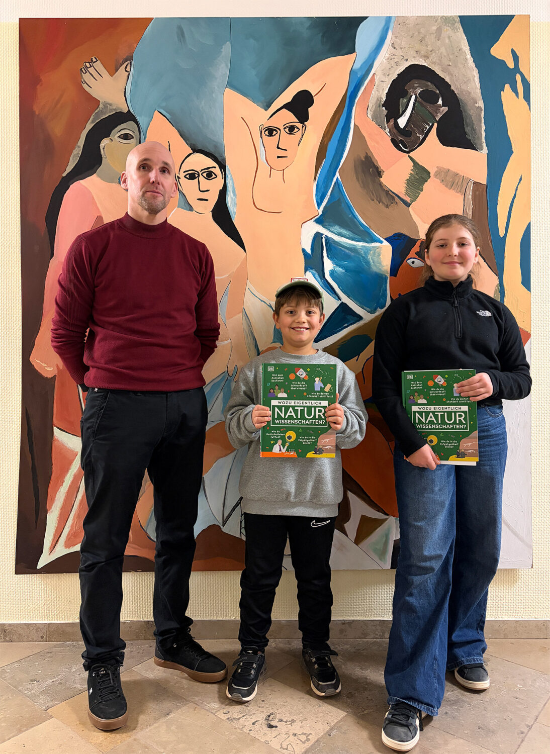 Ein Mann und zwei Kinder, die Bücher in der Hand halten, stehen vor einem farbenfrohen abstrakten Gemälde mit menschlichen Figuren.