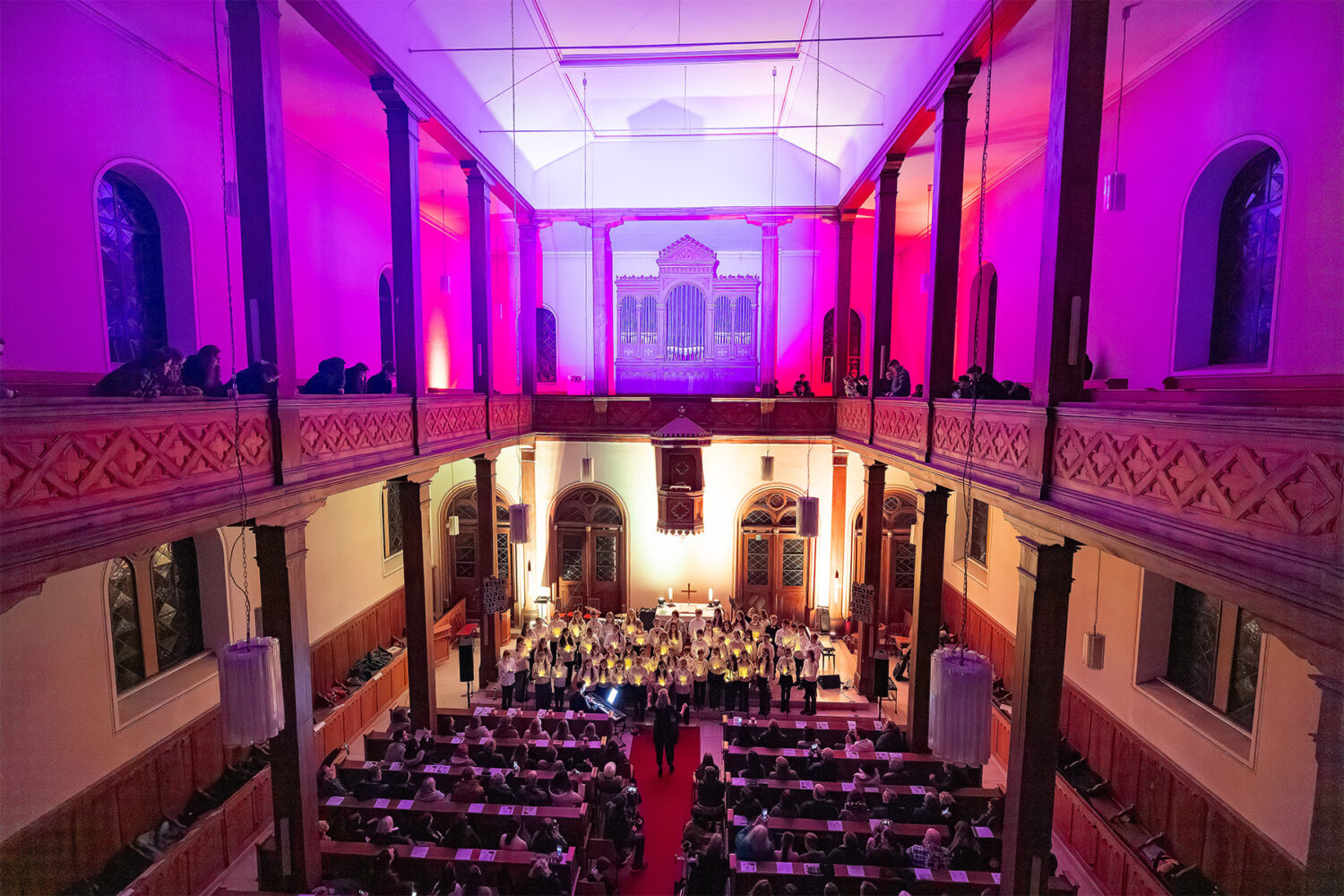 Ein Konzert oder eine Aufführung in einem großen Saal mit farbenfroher violetter Beleuchtung und einem großen Publikum, das zusieht.