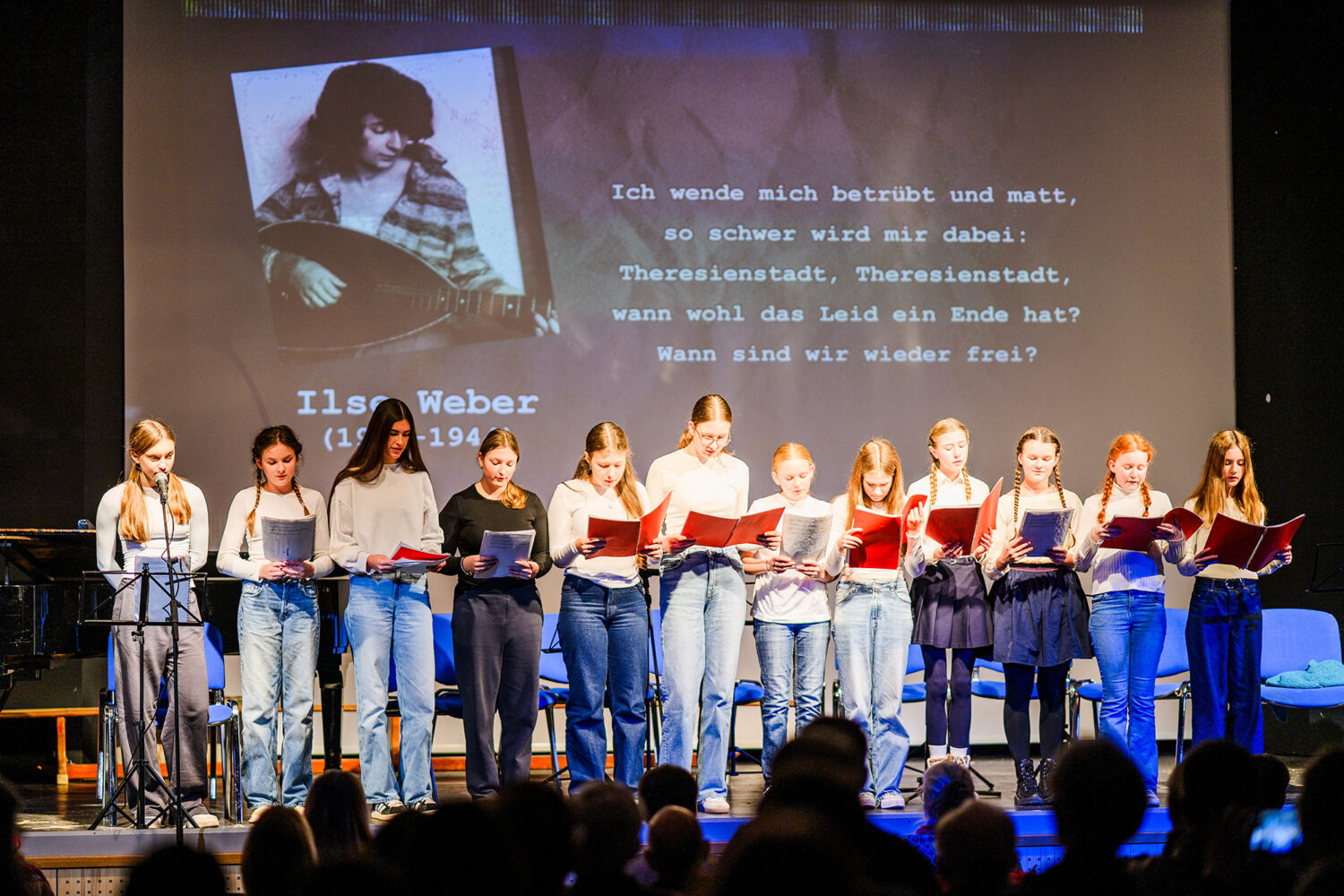 Zwölf Mädchen stehen auf der Bühne und lesen aus roten Mappen vor, hinter ihnen ein projiziertes Foto und ein deutscher Text.