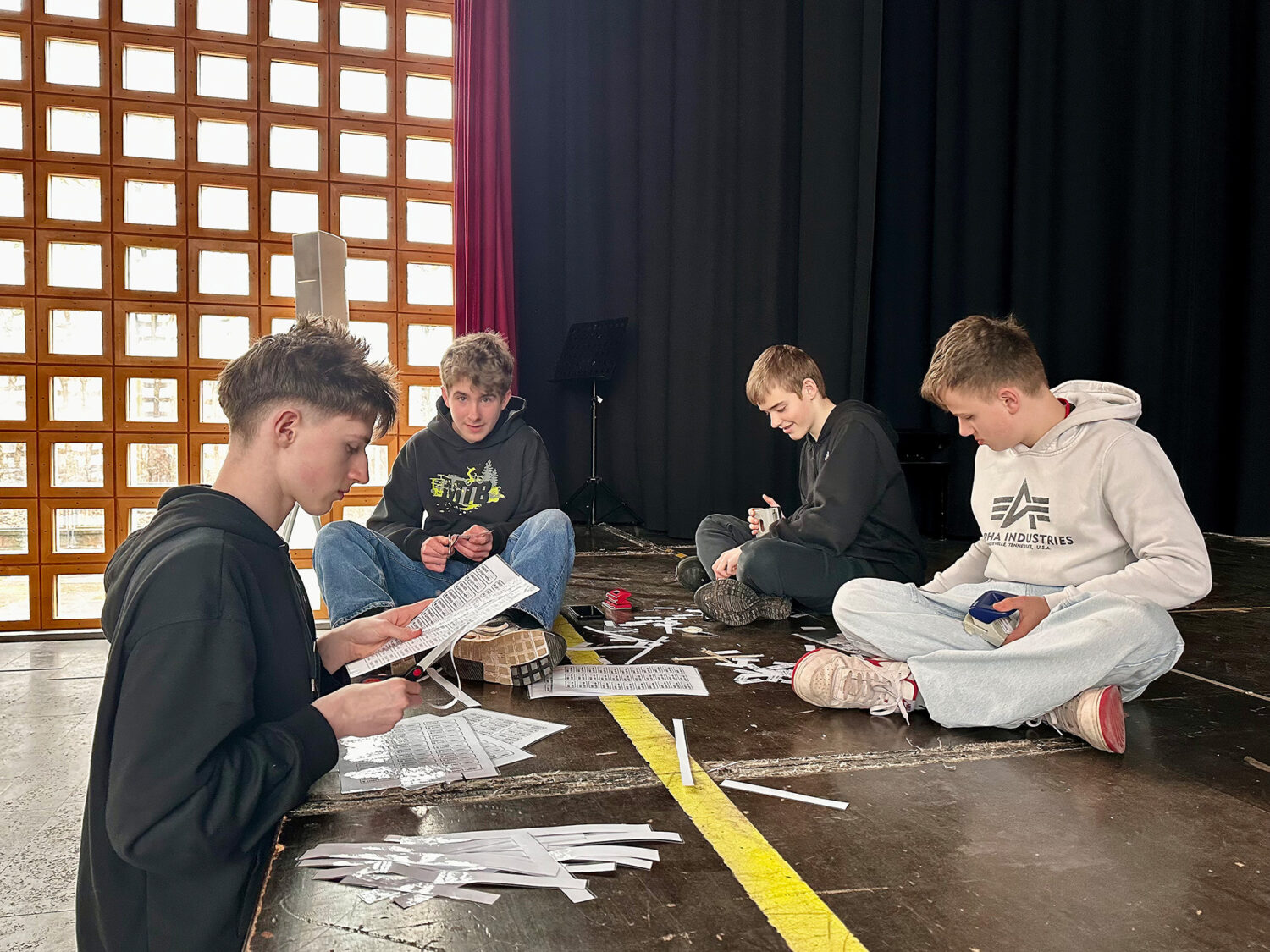 Vier Jungen sitzen auf einem Bühnenboden und schneiden und sortieren in einem gut beleuchteten Raum Papierstreifen.