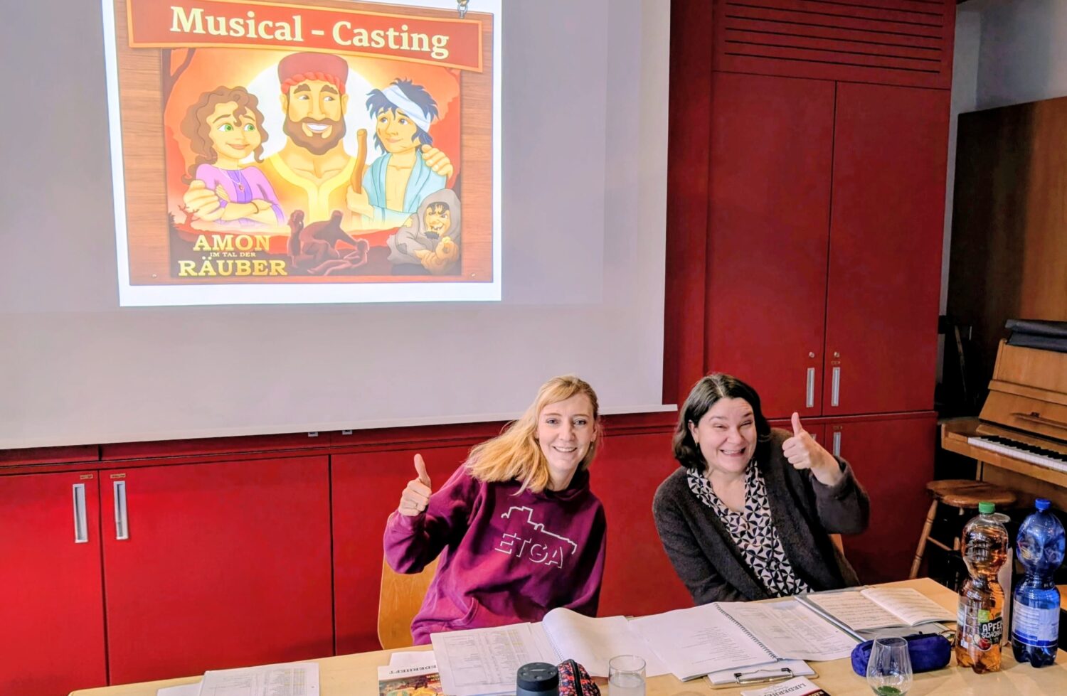 Zwei Frauen sitzen an einem Tisch, lächeln und zeigen die Daumen nach oben bei einem Musical-Casting mit einem Plakat im Hintergrund.