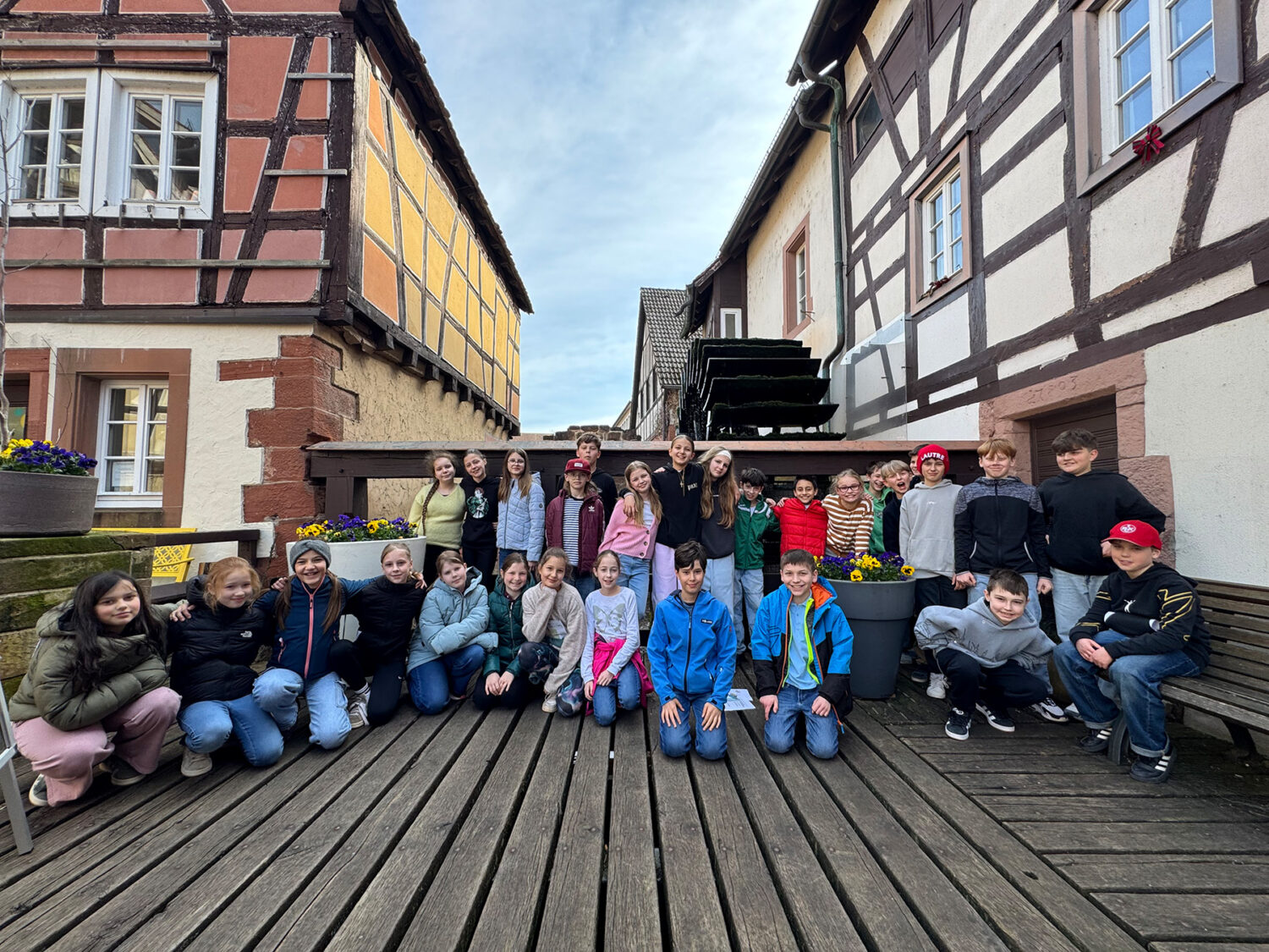 Eine Gruppe von Kindern posiert draußen zwischen zwei Fachwerkhäusern auf einem Holzdeck zusammen.