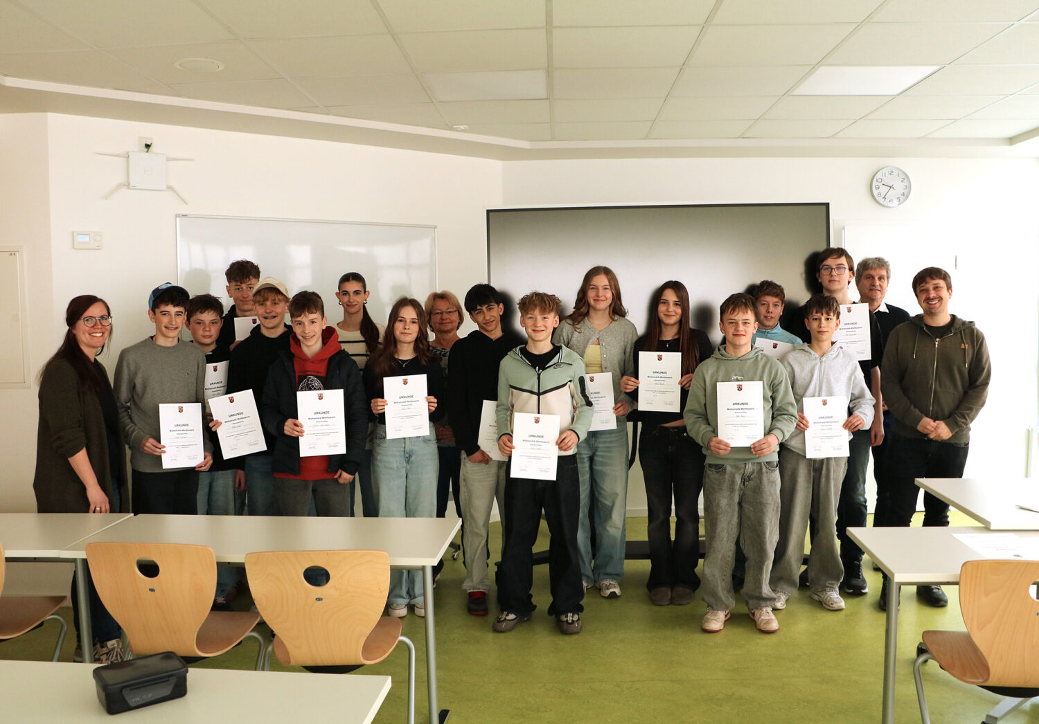 Eine Gruppe Jugendlicher und zwei Erwachsene stehen in einem Klassenzimmer, halten Urkunden in der Hand und lächeln in die Kamera.