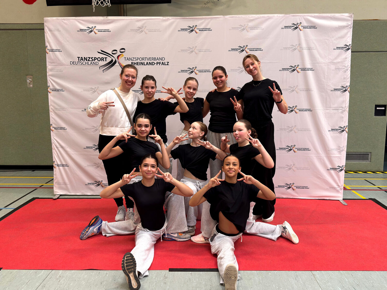Neun Mädchen in schwarzen und weißen Outfits posieren auf einem roten Teppich mit Friedenszeichen vor dem Hintergrund einer Tanzveranstaltung.