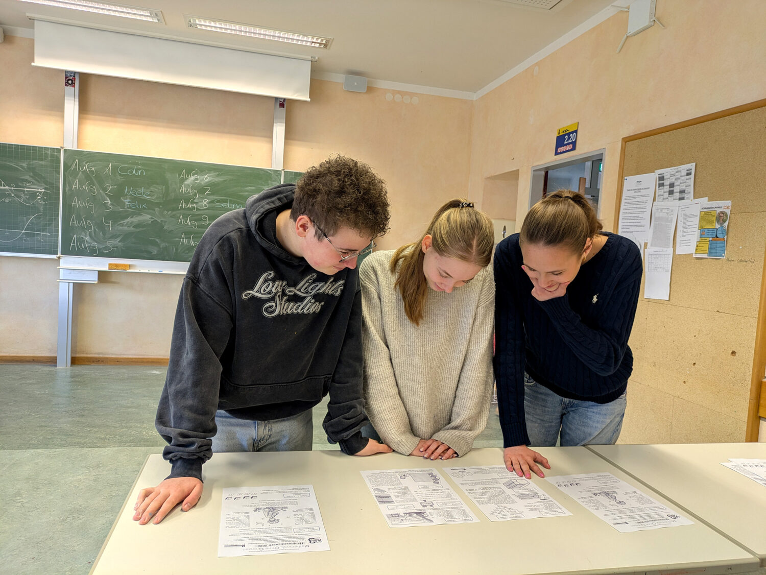 Drei Schüler stehen an einem Tisch in einem Klassenzimmer und betrachten aufmerksam die vor ihnen ausgebreiteten Papiere.