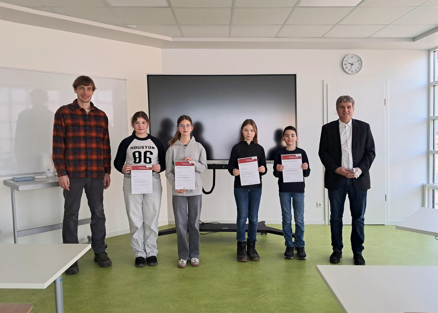 Fünf Personen posieren in einem Klassenzimmer; vier Schüler halten Zeugnisse, zwei Erwachsene stehen neben ihnen.