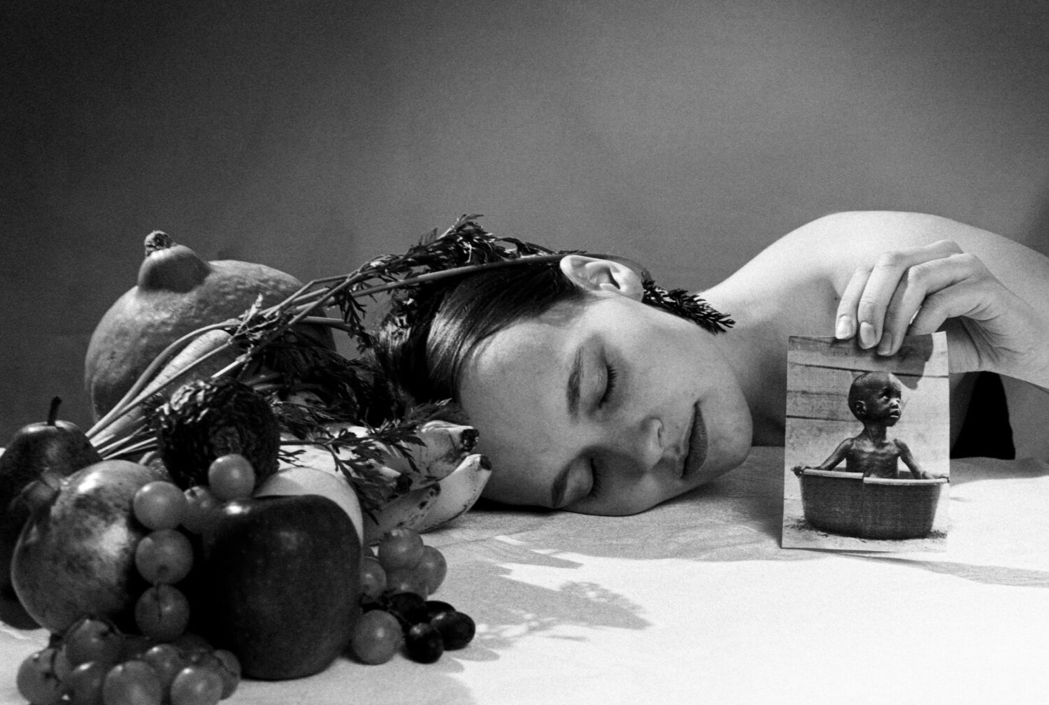 Eine Frau stützt ihren Kopf auf einen Tisch mit Obst und hält ein Foto eines Kindes in einer kleinen Wanne.