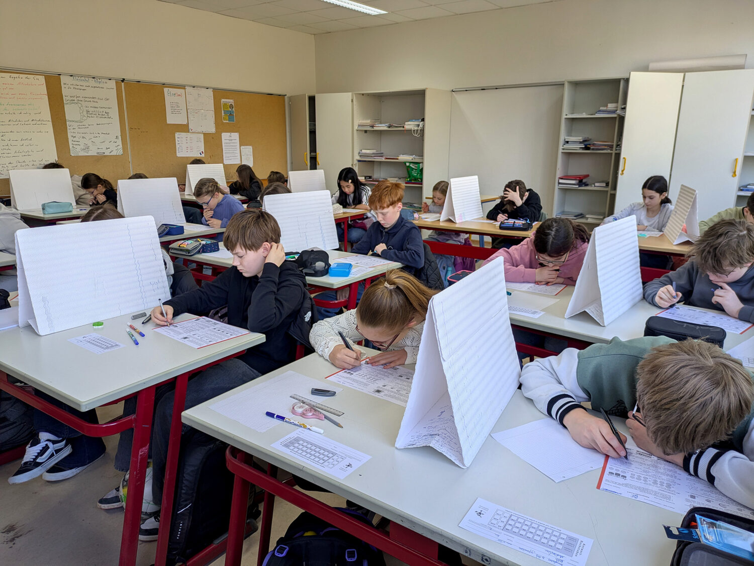 Die Schülerinnen und Schüler sitzen an Schreibtischen mit Trennwänden, konzentriert auf das Schreiben, in einem Klassenzimmer mit Whiteboards und Regalen.