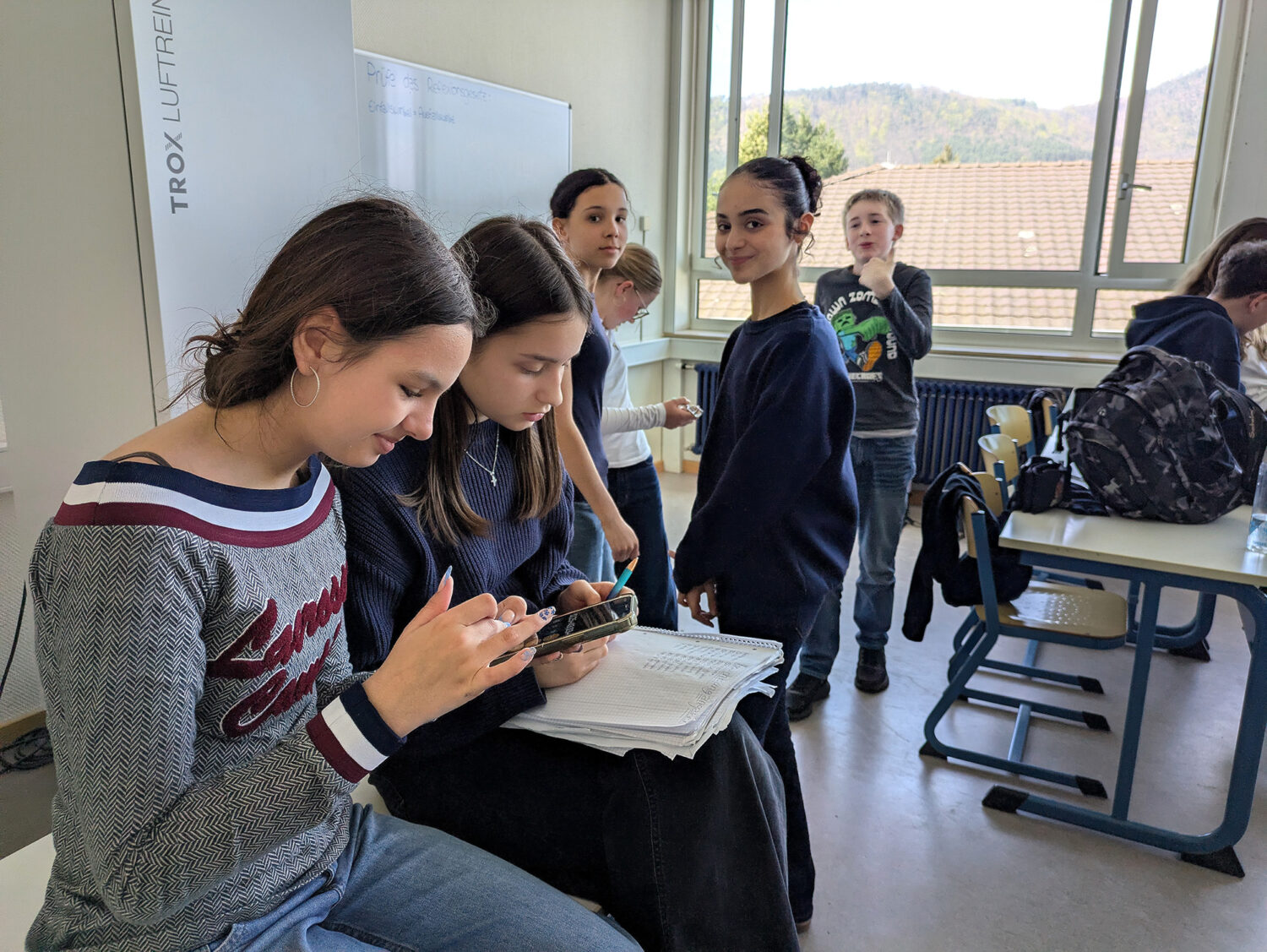 Mehrere Schüler versammeln sich in einem Klassenzimmer; zwei Mädchen konzentrieren sich auf ein Telefon, während andere im Hintergrund stehen und sich unterhalten.