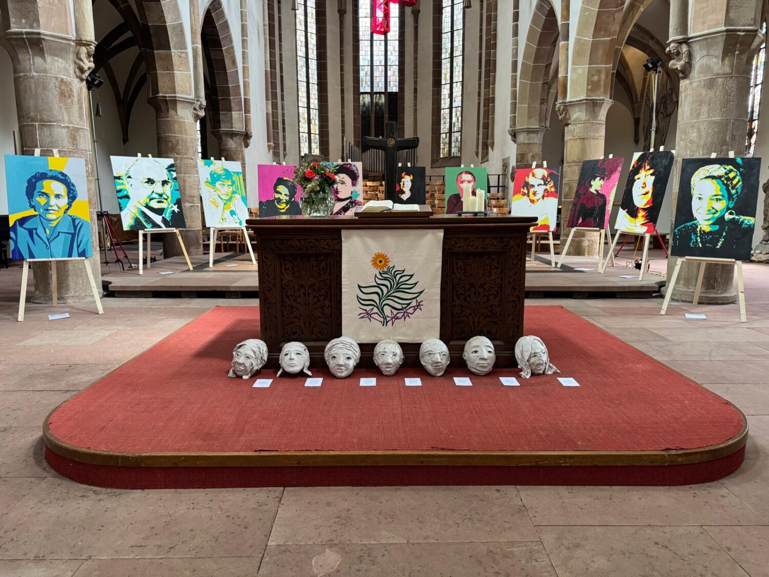 Kunstausstellung in einer Kirche mit bunten Porträts auf Staffeleien und skulptierten Köpfen vor einem Altar.