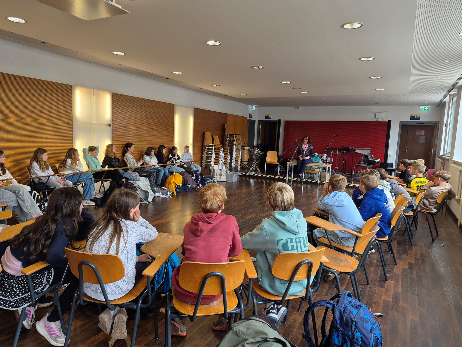 Eine Gruppe von Schülern sitzt in einem hellen Klassenzimmer mit Holzfußboden im Kreis vor dem Lehrer.