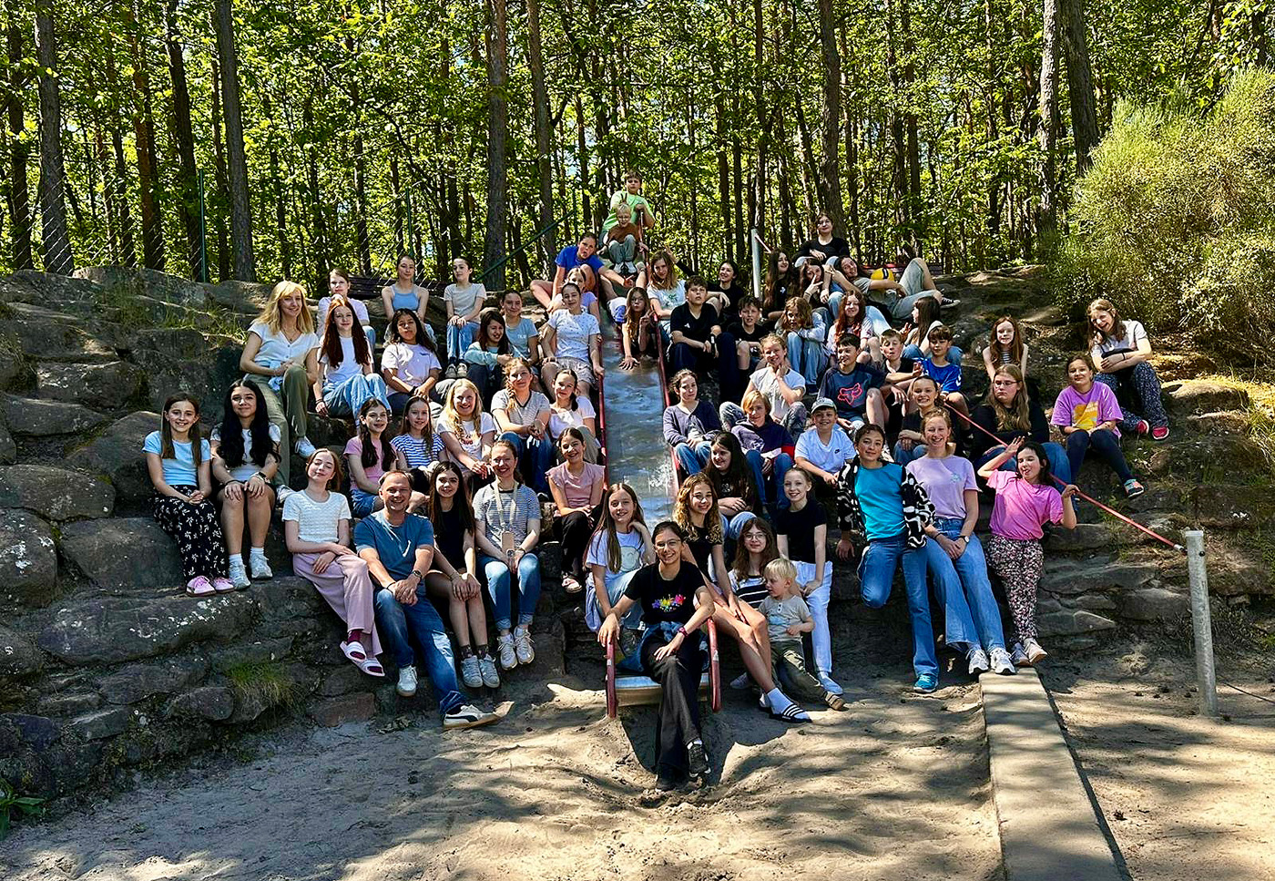 Eine große Gruppe von Jugendlichen und Erwachsenen sitzt auf Steinstufen in einem sonnigen, bewaldeten Außenbereich und lächelt in die Kamera.
