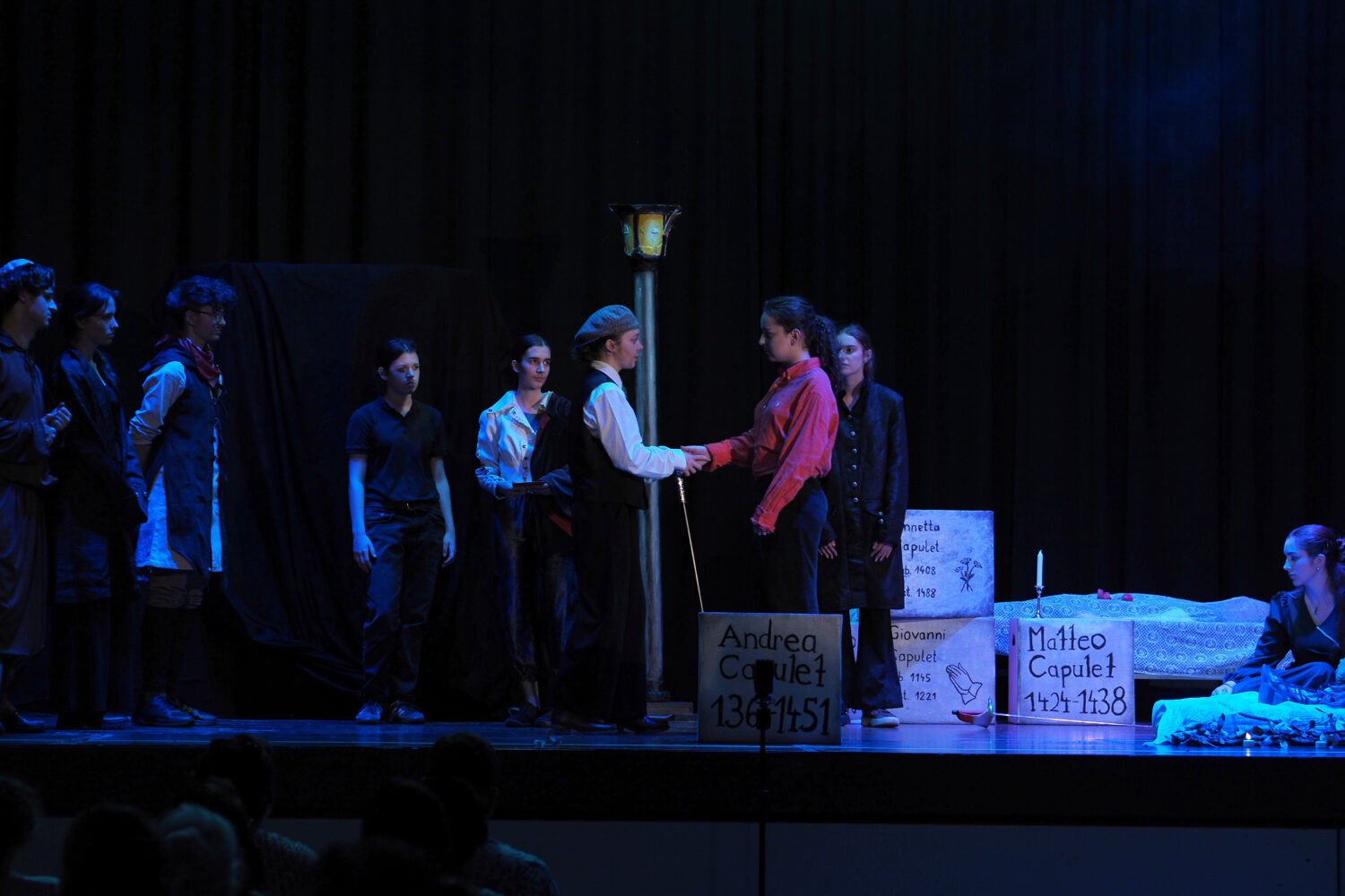Eine Gruppe von Schauspielern spielt auf der Bühne eine dramatische Szene, wobei Grabsteine und gedämpftes Licht eine düstere Stimmung erzeugen.