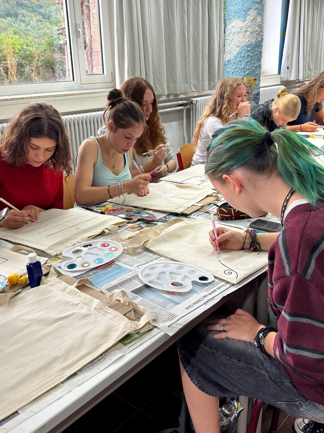 Eine Gruppe junger Leute sitzt an einem Tisch und bemalt Stofftaschen mit Pinseln und Acrylfarben.