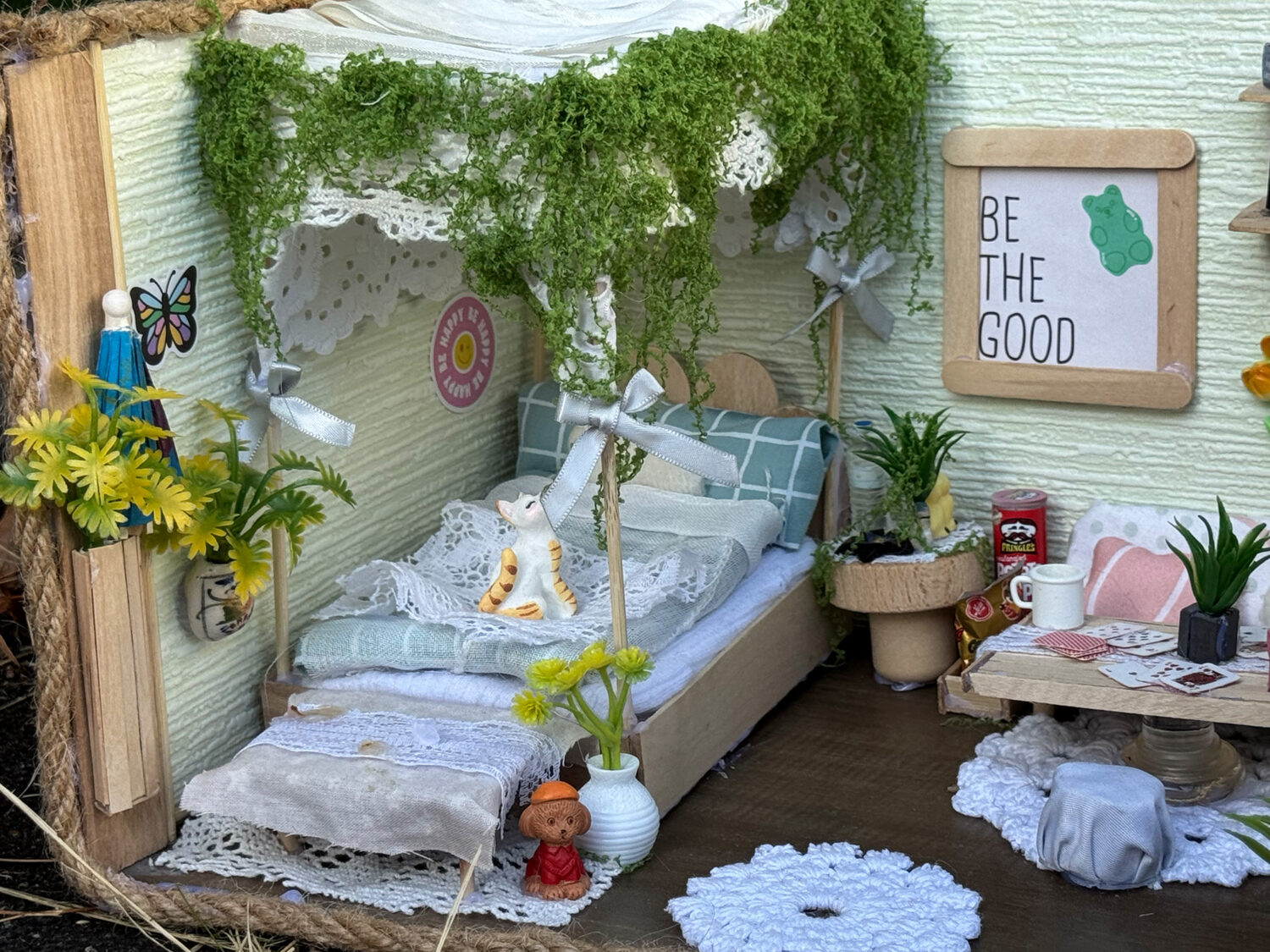 Ein gemütliches Miniatur-Schlafzimmer mit Pflanzen, einem Bett mit Spitzenhimmel, winzigen Dekoelementen und einem Wandschild mit der Aufschrift „Be The Good“.