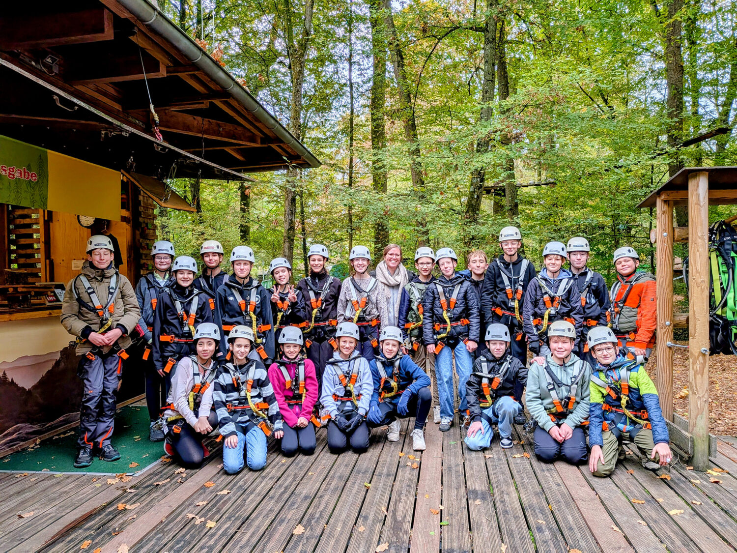 Eine große Gruppe von Kindern und ein Erwachsener, alle mit Klettergurten und Helmen ausgestattet, stehen in einem Wald.